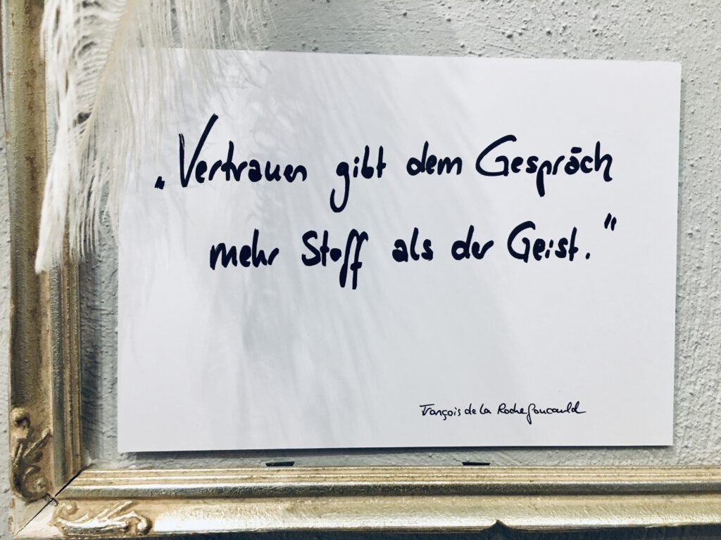 In einem goldenen Rahmen hängt ein handbeschriebenes Blatt mit einem Zitat von François de La Rochefoucauld. Dort steht „Vertrauen gibt dem Gespräch mehr Stoff als der Geist.“