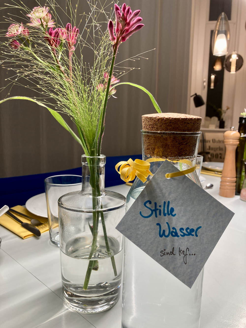 Auf einem Tisch stehen Blumen, ein Gedeck sowie eine Wasserflasche, an der ein Kärtchen hängt mit der Aufschrift „ Stille Wasser ... sind tief“.