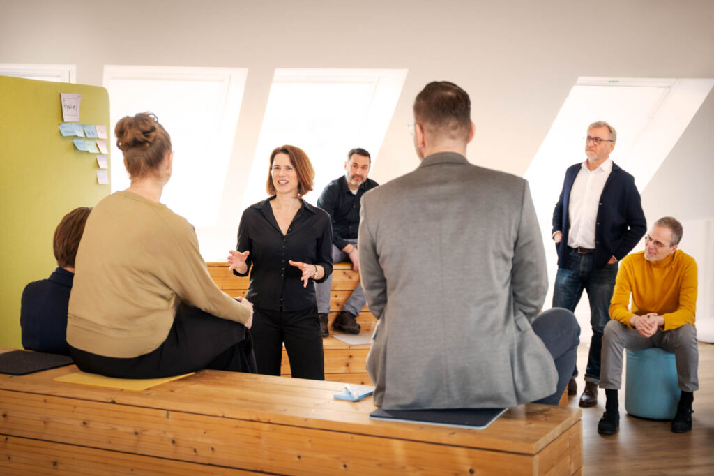 Zwei Menschen sitzen mit dem Rücken zum Betrachter auf einer Holzbank, Anna Brandes steht vor ihnen und moderiert. Im Hintergrund sind weitere Workshop-Teilnehmende zu sehen.
