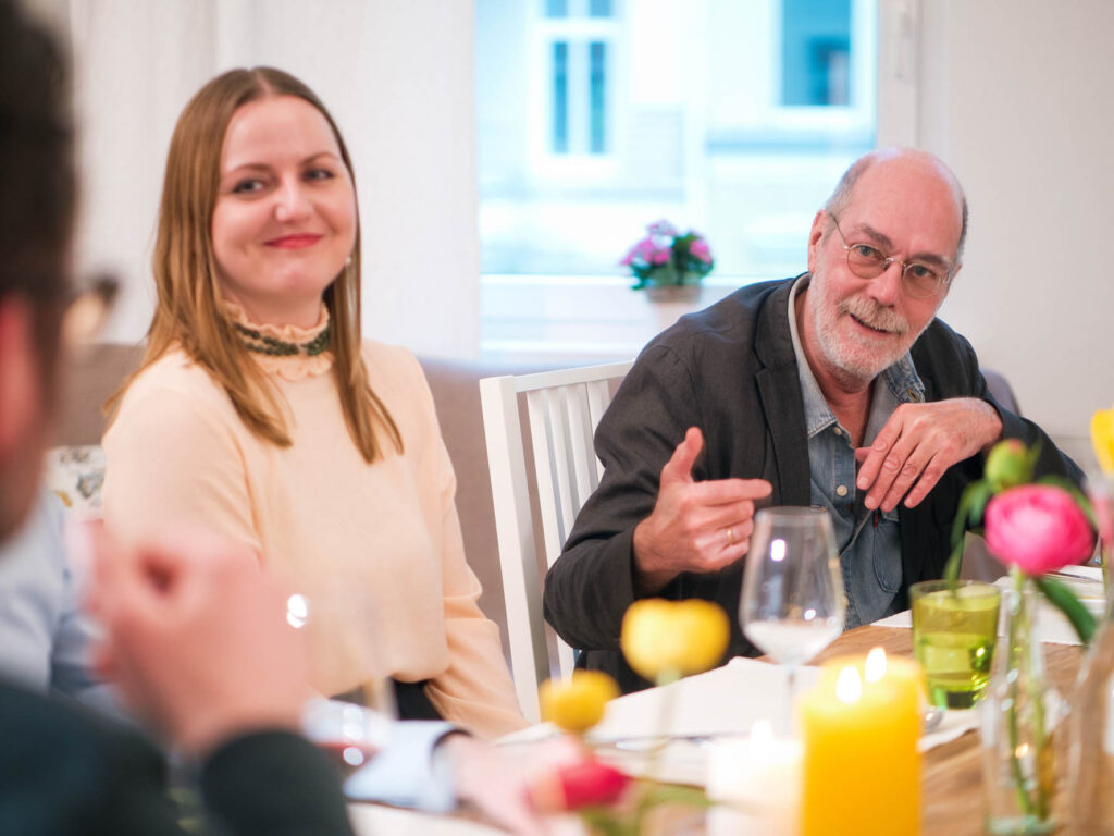 Eine Gesprächsszene am bunt gedeckten Tisch. Eine Frau in creme-farbener Bluse und ein gestikulierender Mann mit grauen Haaren blicken beide in Richtung eines weiteren Mannes, der am Kopfende des Tisches sitzt.