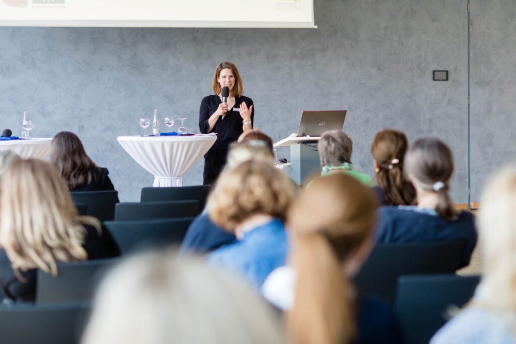 Im Vordergrund des Bildes sitzen mehrere Frauen in Stuhlreihen verteilt und blicken nach vorne zur Bühne, auf der Anna Brandes mit Mikrofon in der Hand steht und moderiert.