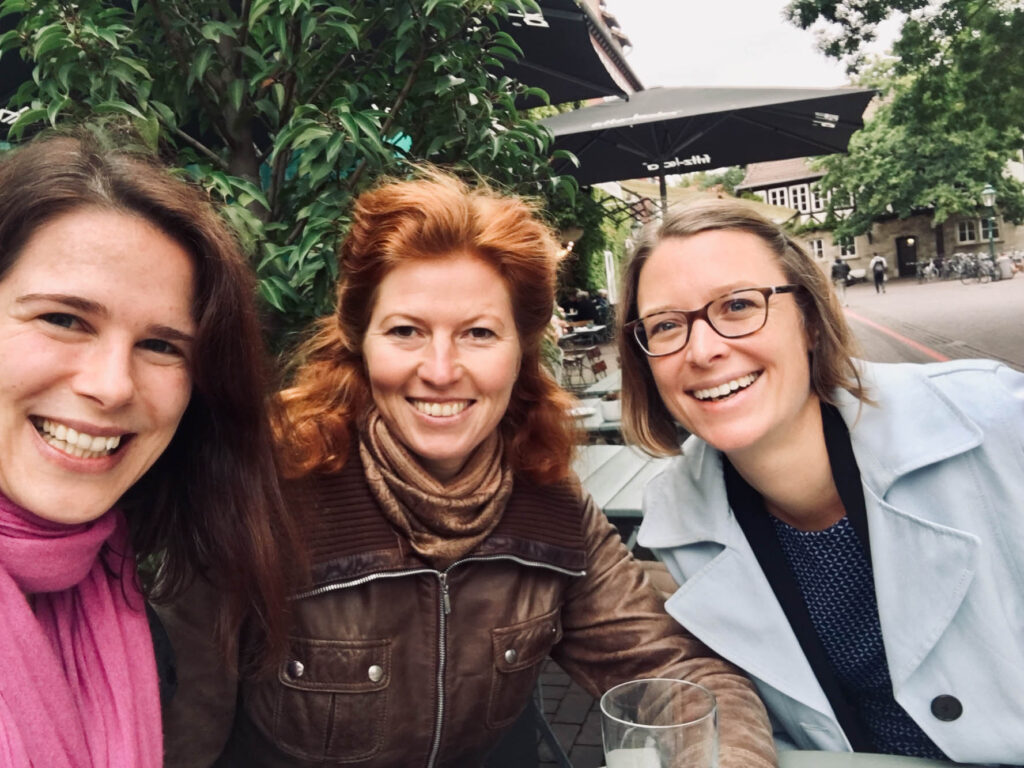 Eine Selfie-Aufnahme von Anna Brandes und zwei Lunch-Connection-Teilnehmerinnen, draußen vor einem grünen Baum und Lokal im Hintergrund. Alle lachen.