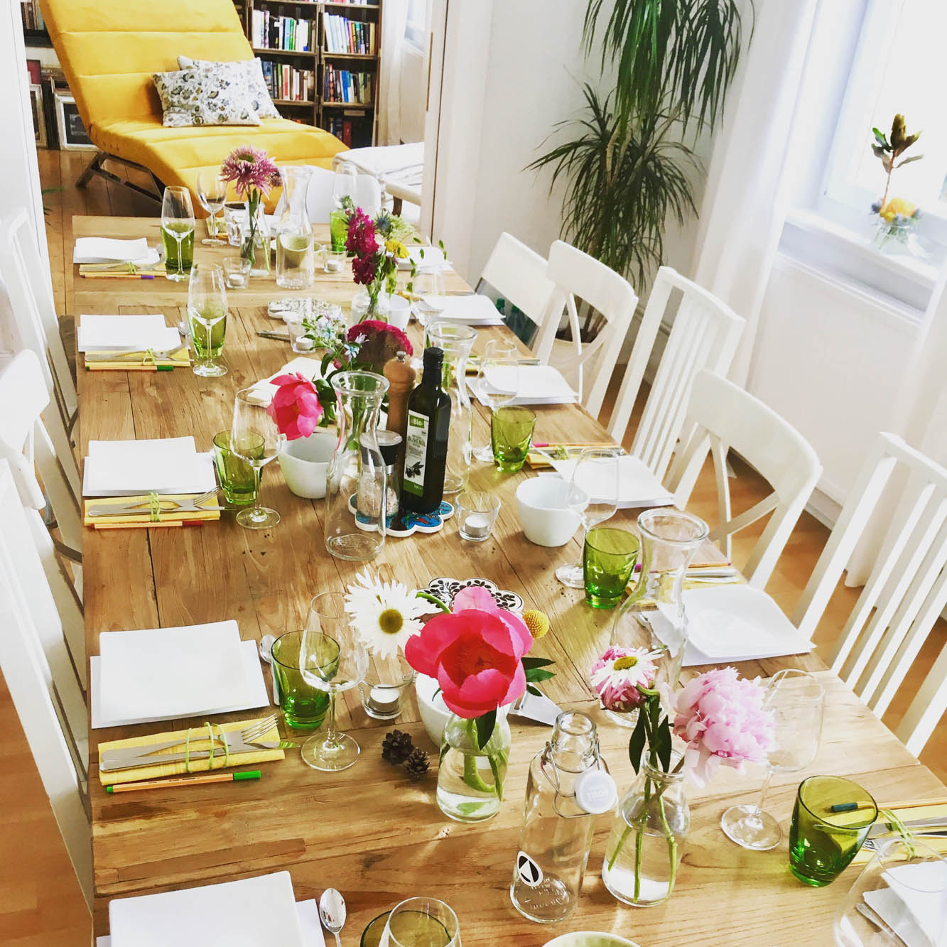 Ein langer Holztisch mit weißem Geschirr für 11 Personen und bunten Blumen. Im Hintergrund steht eine gelbe Sofaliege. Die Atmosphäre wirkt einladend und warm.