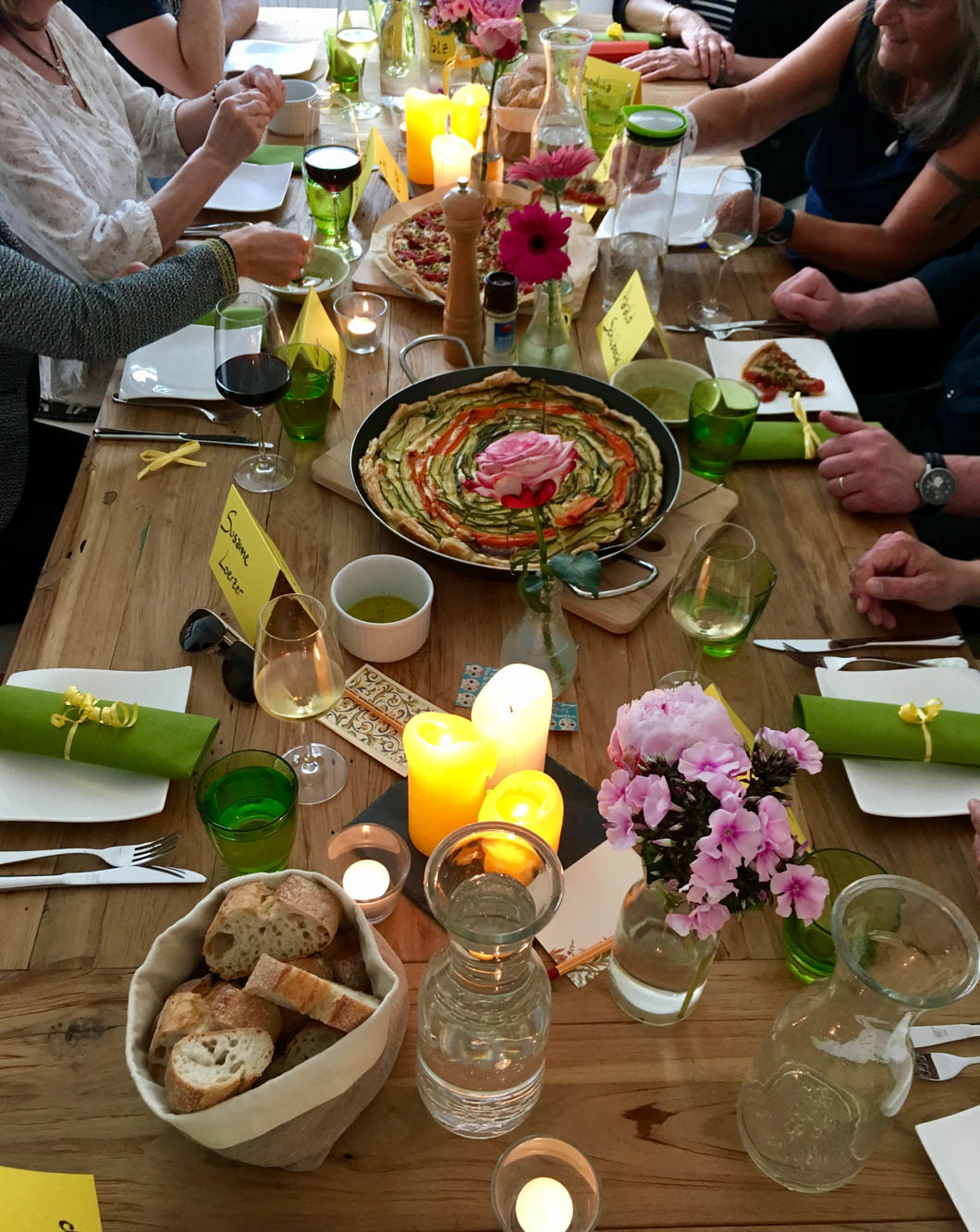 Ein reich gedeckter Holztisch mit bunten Quiches, Baguette, Gedecken, leuchtenden Kerzen und Blumen. Drumherum sitzen Menschen, die sich an den Köstlichkeiten bedienen.
