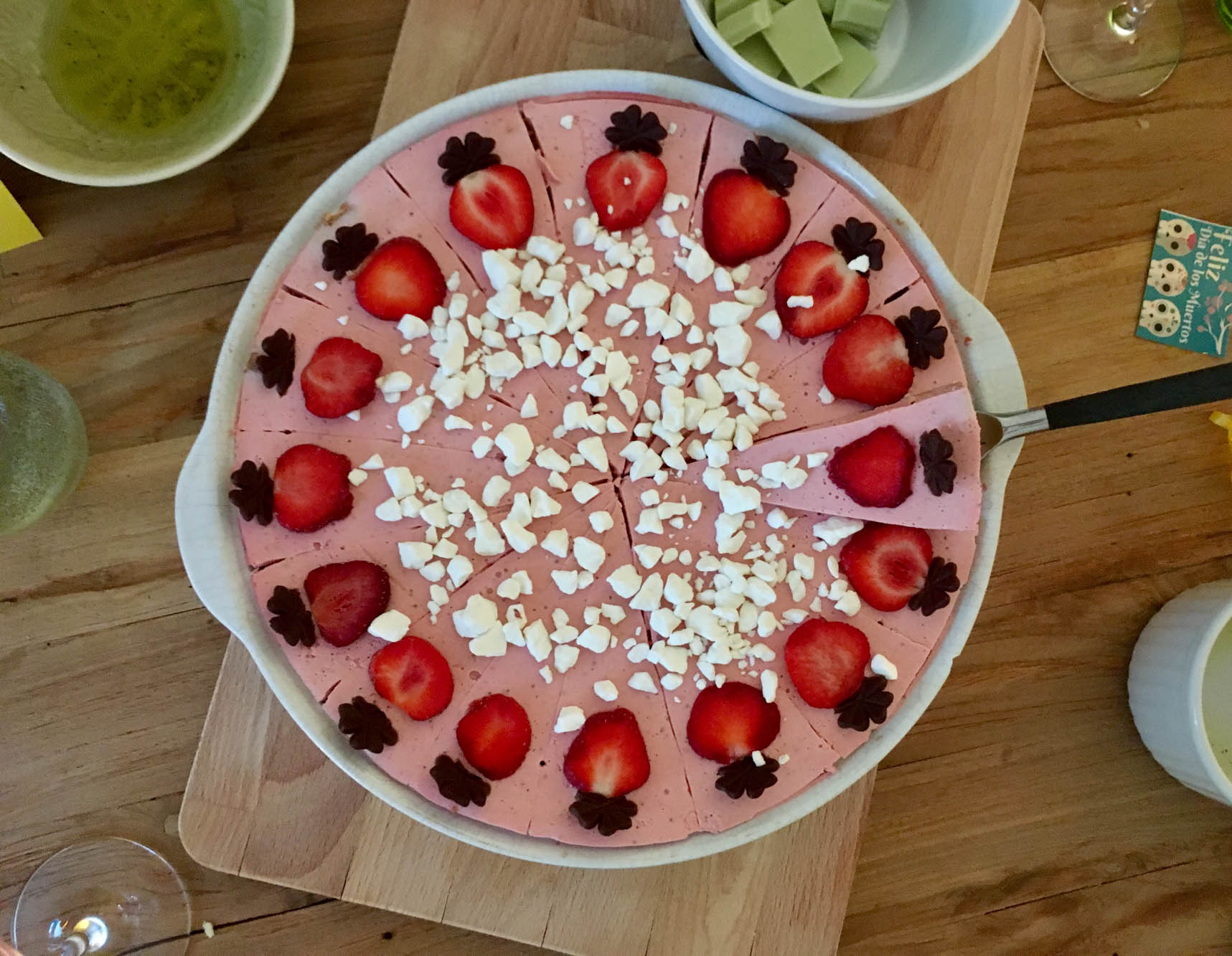 Ein rosa-farbener Erdbeer-Kuchen von oben aufgenommen, in einer runden Form. Unter einem Kuchenstück befindet sich ein Tortenheber – bereit, gegriffen zu werden.