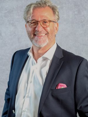 Uwe Rissiek Profilbild von Uwe Rissiek. Er trägt ein weißes Hemd und dunkles Sacko mit rosa Einstecktuch und blickt offen lachend in die Kamera.